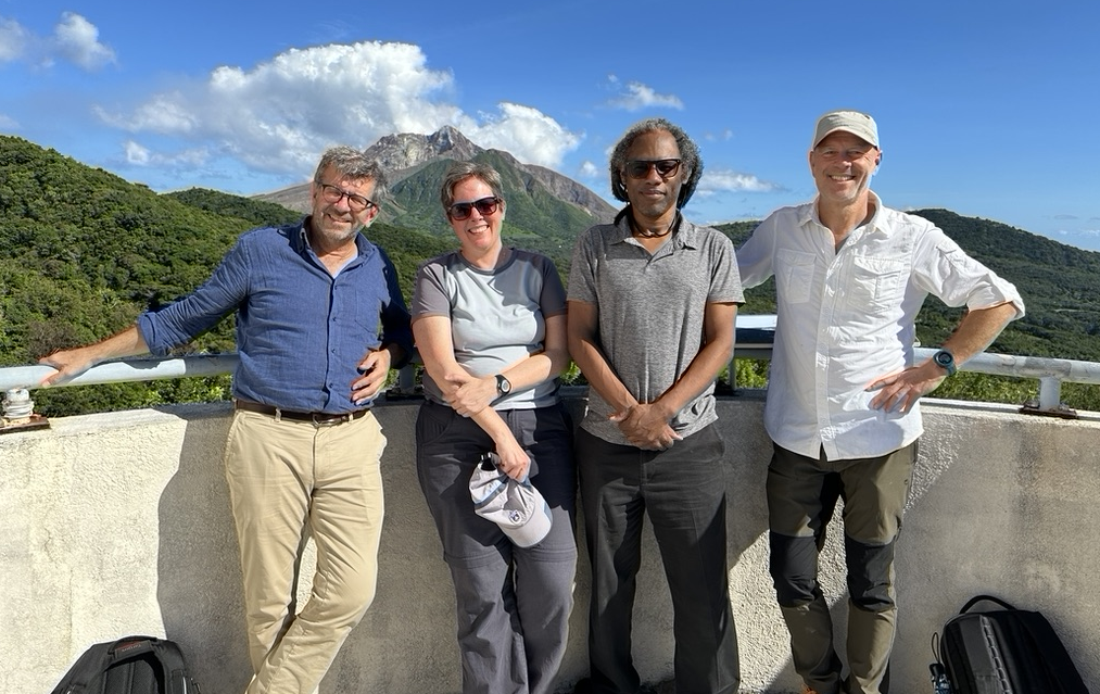 A group of three men and a woman stand on a observation point. In the background are forested hills and an active (but not erupting) volcano. 