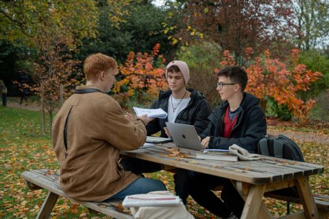 Photo of three students sat around a picnic table surrounded by autumn trees