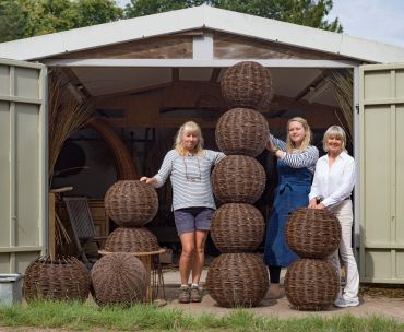 Three white women stand in front of a workshop with parts of their installation - balls made of willow