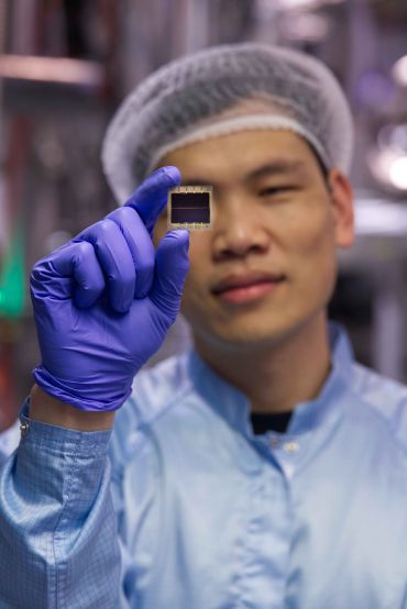 An Asian researcher wearing a lab coat, hairnet, and latex gloves holds in his hand a very thin material that looks like a computer chip.  