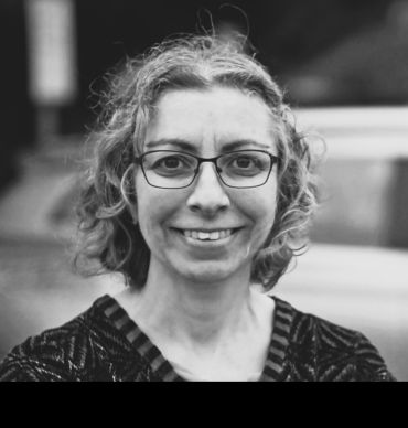 A portrait black and white photograph of Dr Hannah Budnitz, a lady with shoulder-length hair wearing glasses.