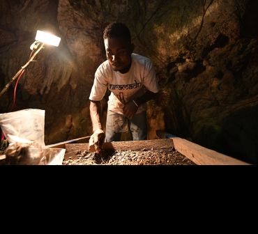 A man stands in a cave, reaching into a square sieve filled with debris. In his hand is a small zip-lock bag containing a few fragments. He is illuminated from above by a lamp. 