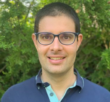 Mark Mezei, a white man with short dark hair and glasses, wearing a dark blue polo shirt. He stands outside, by a hedge.