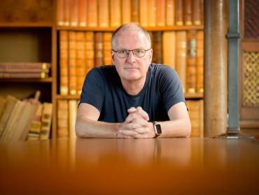 Professor Michael Wooldridge, a white man with glasses, sitting at a table in front of a bookshelf filled with old books.