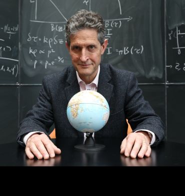 A portrait photograph of a white man wearing a dark suit, seated at the table with a 3D Globe model in front of him. In the background is a blackboard with mathematical equations scribbled on it.