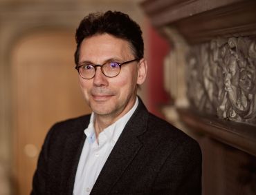 Portrait of Sam Fankhauser, a middle aged man with dark hair wearing a black suit and white shirt, standing by a fireplace.