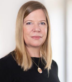 Dr Paula Sheppard, a middle-aged white lady with shoulder length blonde hair. She wears a black top with a golden pendant, and stands in front of a white wall.