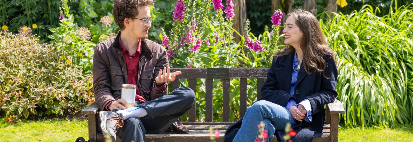 Two students sit at opposite ends of a bench facing each other. They are talking and smiling.