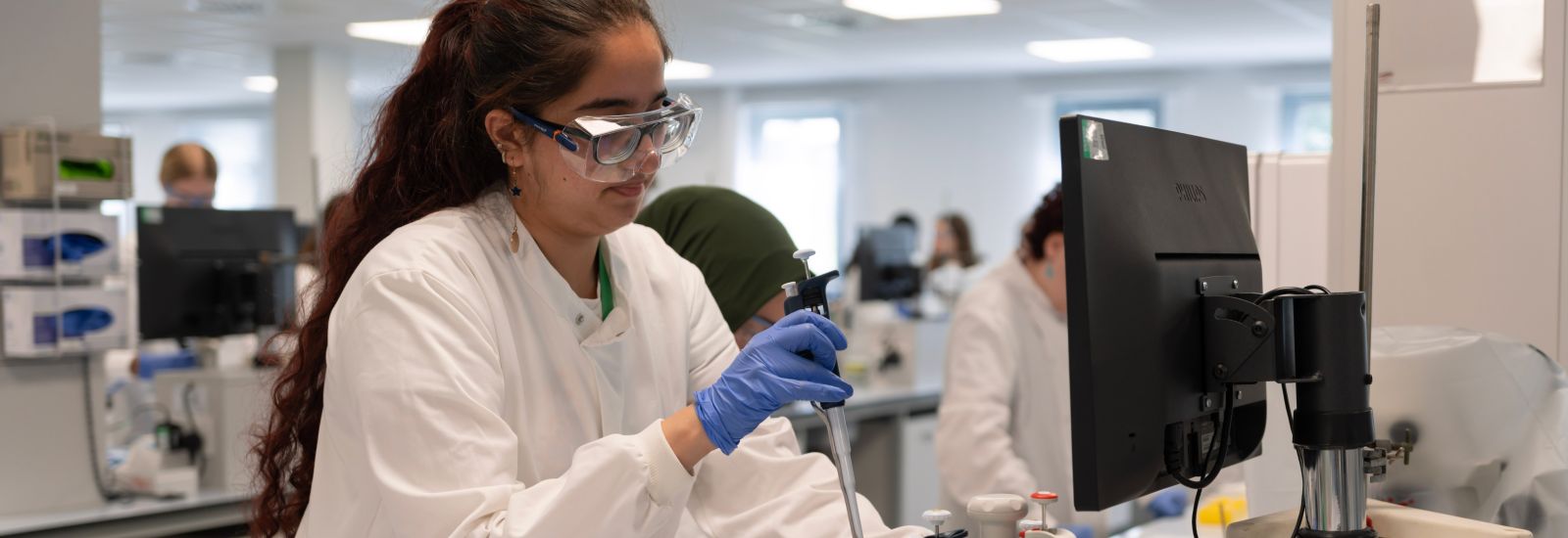 A student in a lab wearing a lab coat, goggles and gloves carries out an experiment.