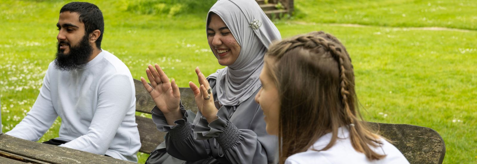 A group of students sits around a wooden table on a patch of grass. They are laughing, smiling and gesturing at one another.