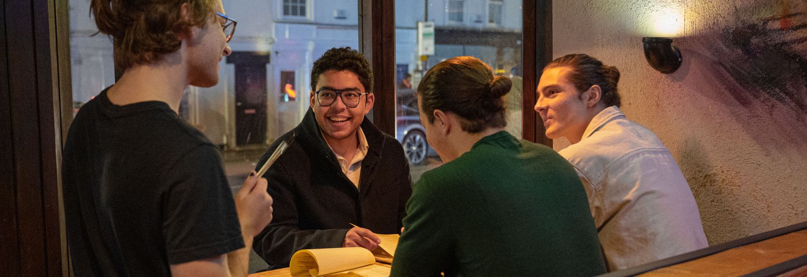 A group of students sit in a restaurant. A waiter takes their orders.