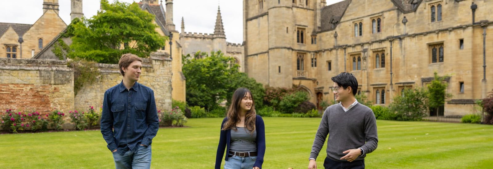 Three students walk across the Magdalen College green. They are smiling and speaking to one another.