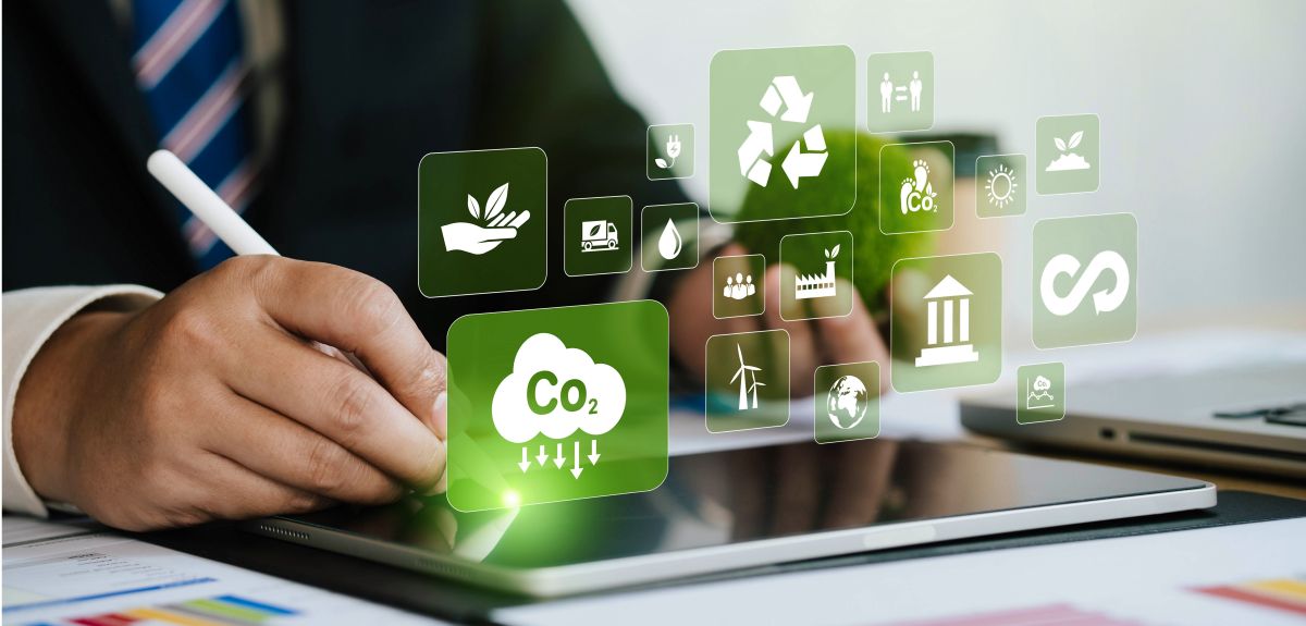 A business person’s hands rest on a desk with a tablet or iPad device and sheets of paper with graphs and charts. Overlaid are icons representing metrics of sustainability, such as carbon emissions, water and recycling.