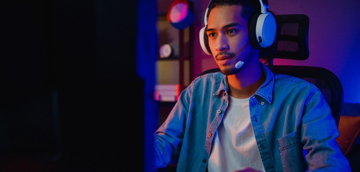 Hombre joven con auriculares y micrófono jugando a videojuegos frente a una pantalla. Está concentrado mientras juega, sentado en una silla de oficina en un ambiente iluminado con luces de colores azul y púrpura, típico de un entorno de gaming.