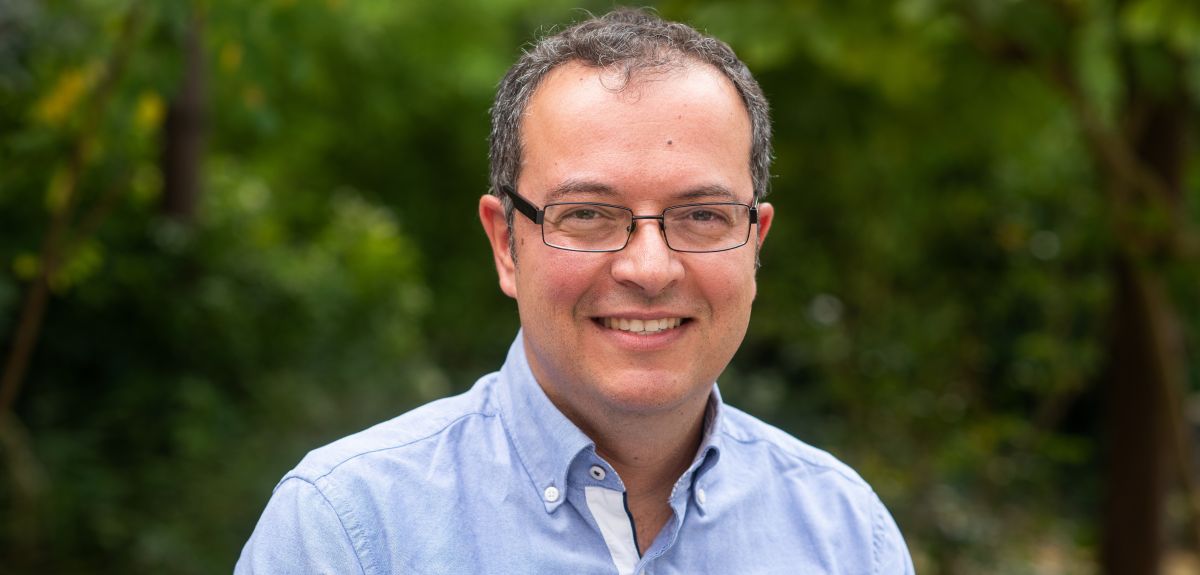 Portrait photograph of Gabriel Stylianides. He is a white man with short brown hair wearing black glasses and a blue shirt. He is outside, in front of an out-of-focus background of trees.