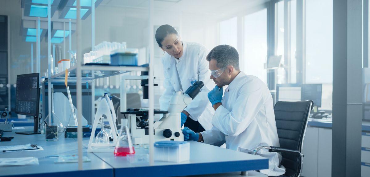 A man and woman wearing lab coats sit at a bench in a laboratory. In front of them, there is a microscope and a range of chemistry apparatus. 