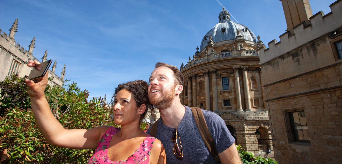 Image of a man and woman talking a selfie with the Radcliffe Camera in the background