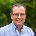 Portrait photograph of Gabriel Stylianides. He is a white man with short brown hair wearing black glasses and a blue shirt. He is outside, in front of an out-of-focus background of trees.