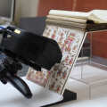 A scanning device analyses a manuscript page help open on a clear Perspex stand. The page features illustrated drawings of human figures in red and gold ink.