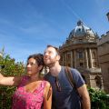 Image of a man and woman talking a selfie with the Radcliffe Camera in the background